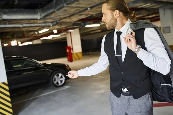Schöner Mann mit Bart in schwarzer Weste mit Autoschlüssel auf Parkplatz, Geschäftskonzept — Stockfoto