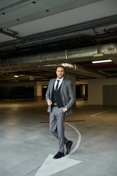 Atractivo hombre de negocios elegante en traje elegante gris posando con la mano en el bolsillo y mirando a la cámara - foto de stock