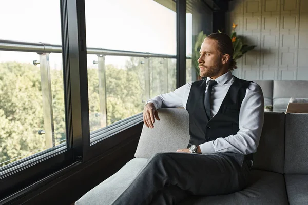 Handsome stylish businessman in elegant smart attire sitting on sofa and looking out of window — Stock Photo
