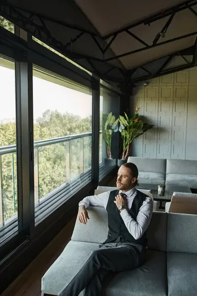 Eleganter Geschäftsmann in schwarzer Weste, der seine Krawatte fixiert, während er auf dem Sofa sitzt und aus dem Fenster schaut — Stockfoto