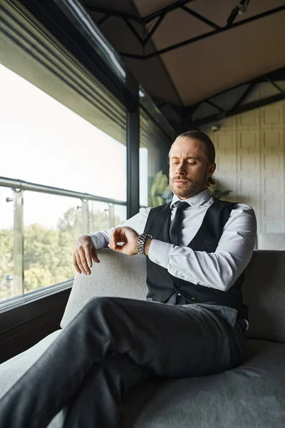 Schöner eleganter Geschäftsmann in schwarzer smarter Weste sitzt auf dem Sofa und schaut auf seine Armbanduhr — Stockfoto