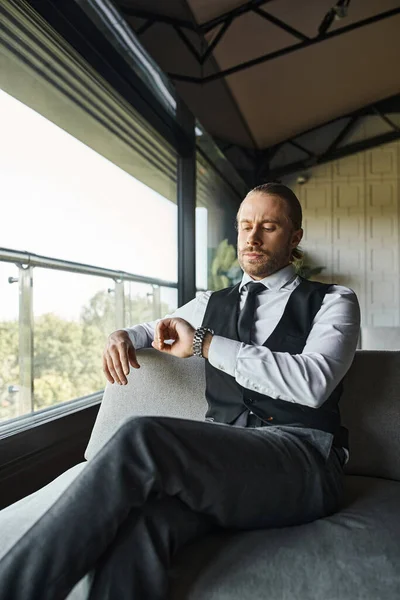 Handsome elegant businessman in black smart vest sitting on sofa and looking on his wristwatch — Stock Photo