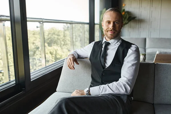 Радостный привлекательный бизнесмен в нарядной одежде сидит на диване и слегка улыбается в камеру — стоковое фото
