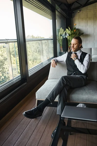Good looking elegant businessman with ponytail in stylish smart black vest sitting on couch — Stock Photo