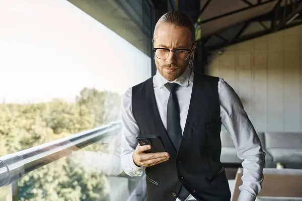 Stylischer Geschäftsmann in schicker schwarzer Weste blickt auf Handy, das am Fenster steht — Stockfoto