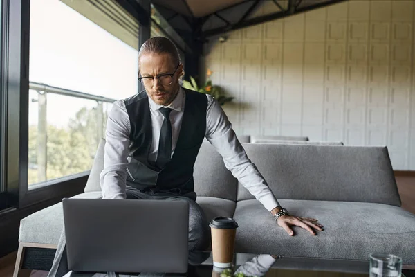 Schöner eleganter Geschäftsmann in schwarzer Weste mit Brille, Blick auf seinen Laptop, Kaffeetasse auf dem Tisch — Stockfoto