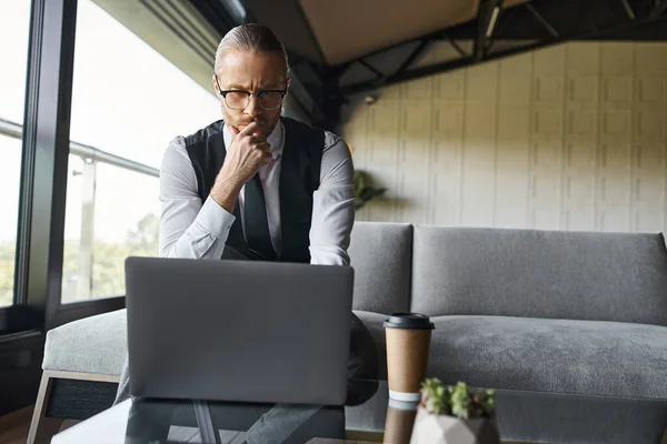 Homem de negócios elegante pensativo em colete preto trabalhando em seu laptop atentamente, xícara de café na mesa — Fotografia de Stock