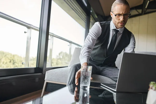 Beau élégant homme d'affaires en tenue intelligente avec cravate travaillant sur ordinateur portable ramassage de verre d'eau — Photo de stock