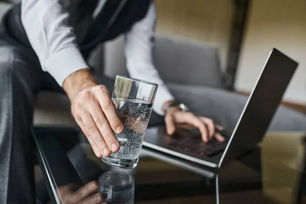 Abgeschnittene Ansicht eines eleganten Geschäftsmannes im grauen schicken Anzug, der ein Glas Wasser nimmt, während er am Laptop arbeitet — Stockfoto