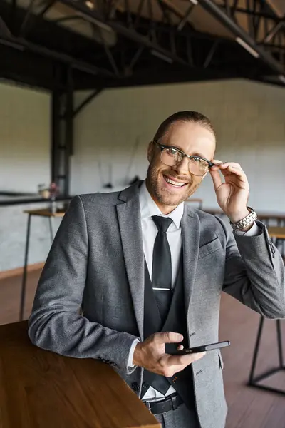 Fröhlicher gutaussehender Geschäftsmann mit Brille im schicken Anzug, der in die Kamera lächelt und sein Handy in der Hand hält — Stockfoto