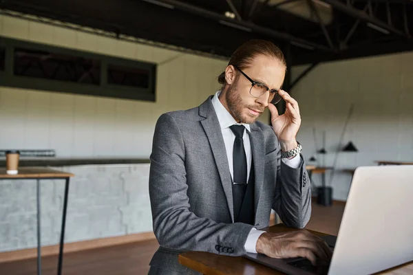 Homem de negócios refinado de boa aparência em terno inteligente cinza trabalhando no laptop com a mão em seus óculos — Fotografia de Stock