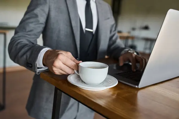 Cropped view of elegant businessman in smart gray suit working on laptop with hand on tea cup — Stock Photo