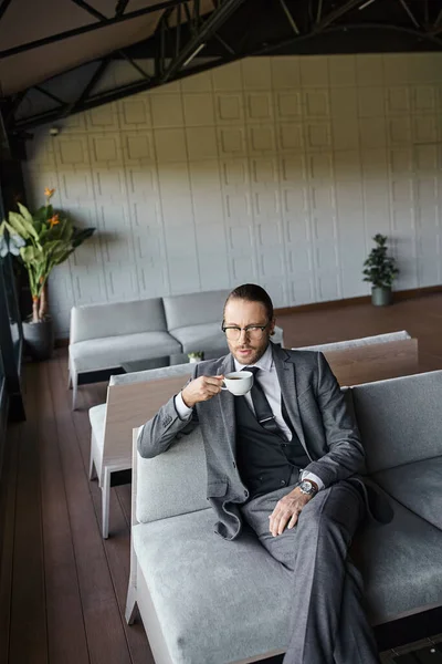 Gut aussehender eleganter Geschäftsmann im grauen schicken Anzug mit Krawatte und Gläsern, die Tee auf dem Sofa trinken — Stockfoto