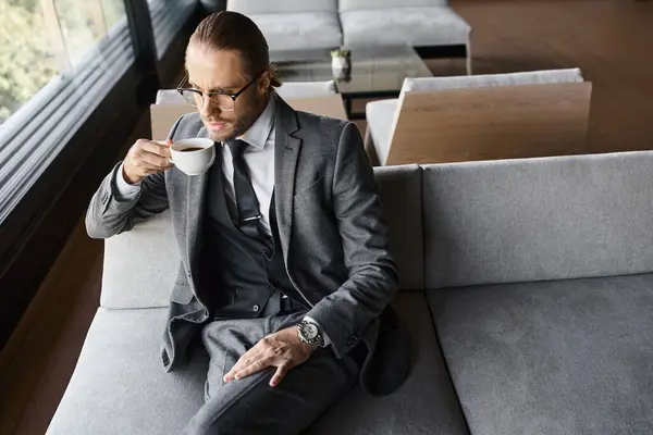 Attraktiver eleganter Mann im smarten grauen Anzug mit Gläsern, die warmen Tee auf dem Sofa trinken, Geschäftskonzept — Stockfoto