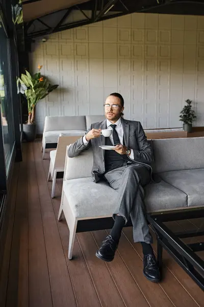 Gut aussehender eleganter Mann im schicken grauen Anzug sitzt auf dem Sofa und trinkt Tee, Geschäftskonzept — Stockfoto