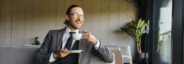 Красивый веселый бизнесмен в элегантном костюме улыбается сидя на диване и пить чай, баннер — стоковое фото
