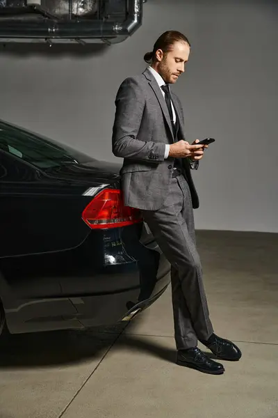 Красивый элегантный бизнесмен в костюме с рыжими волосами, прислонившийся к машине и смотрящий на телефон — стоковое фото
