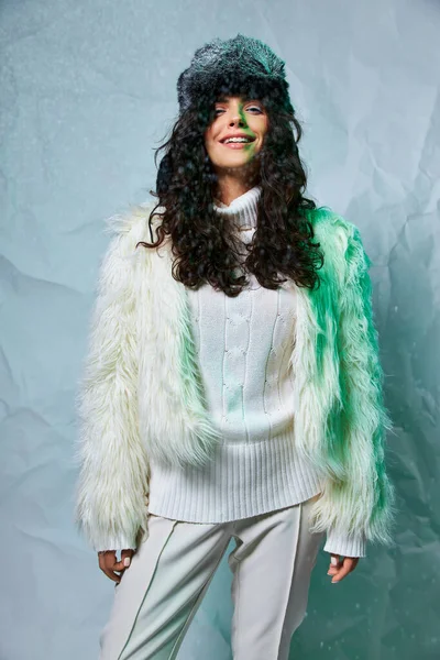 Happy young woman with furry hat, white sweater and faux fur jacket on grey backdrop, winter fashion — Stock Photo