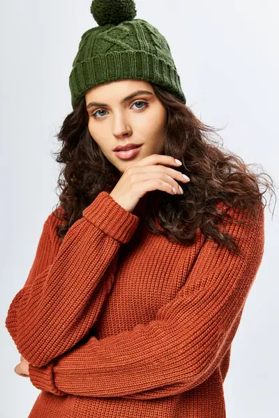 Красивая женщина в шляпе с помпоном и вязаными терракотовыми свитерами позирует на сером фоне — стоковое фото