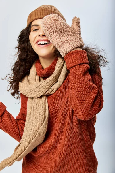 Mulher alegre em chapéu e camisola de terracota de malha cobrindo rosto com mitenes no fundo cinza — Fotografia de Stock