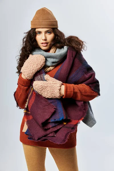 Gemütliche Schichten, lockige junge Frau in mehrschichtiger Kleidung mit warmem Schal und Handschuhen vor grauem Hintergrund — Stockfoto