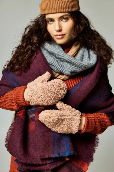 Cozy layers, curly brunette woman in layered clothes with warm scarf and mittens on grey backdrop — Stock Photo