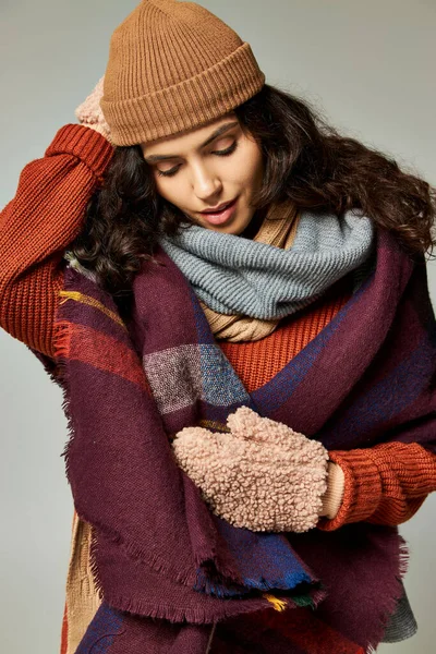 Winter fashion, curly brunette woman in layered clothes with warm scarf and mittens on grey backdrop — Stock Photo