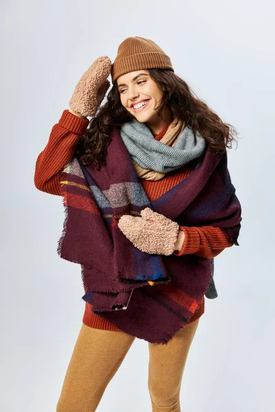 Wintermode, glücklich gelockte Frau in mehrlagiger Kleidung mit warmem Schal und Handschuhen vor grauem Hintergrund — Stockfoto