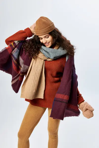 Winter style, cheerful young woman in layered clothing, knitted hat and mittens on grey backdrop — Stock Photo