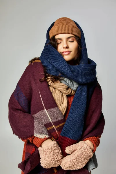 Stile invernale, giovane donna in abiti a strati, cappello a maglia e sciarpe in posa su sfondo grigio — Foto stock