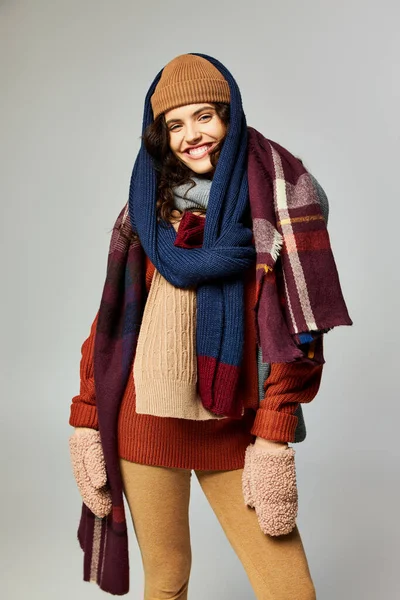 Wintermode, fröhliches Model in mehrschichtiger Kleidung, warme Mütze und Schals posieren vor grauem Hintergrund — Stockfoto