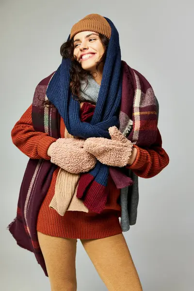 Moda de invierno, modelo alegre en capas de ropa, sombrero caliente y bufandas posando sobre fondo gris - foto de stock