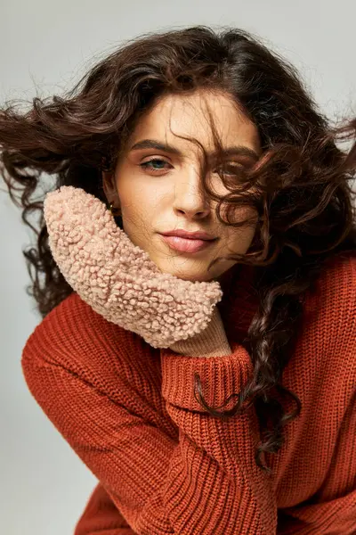 Viento soplando en la cara de la mujer morena rizada en suéter de terracota y mitones, moda de invierno - foto de stock