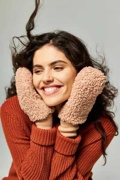 Cheerful and curly brunette woman in terracotta sweater and mittens posing on grey, winter fashion — Stock Photo