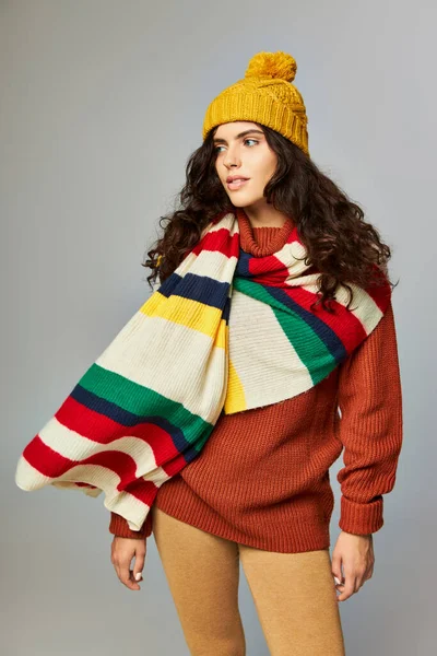 Curly woman in knitted bobble hat and sweater with stripped scarf on top posing on grey backdrop — Stock Photo