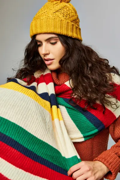 Красивая женщина в вязаной шапке и свитере с раздетым шарфом сверху позирует на сером фоне — стоковое фото