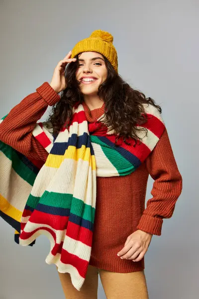 Счастливая молодая женщина в вязаной шапке и свитере с раздетым шарфом позирует на сером фоне — стоковое фото