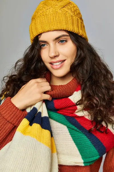 Веселая женщина в зимней шляпе и свитере с раздетым шарфом, позирующая на сером фоне — стоковое фото