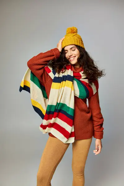 Mulher positiva no inverno bobble chapéu e suéter com cachecol despojado posando no fundo cinza — Fotografia de Stock