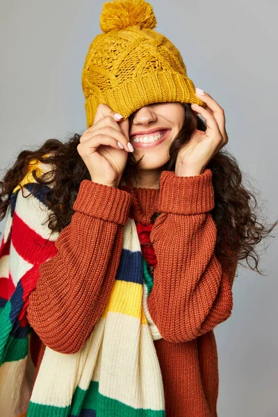 Щаслива жінка в теплому капелюсі і светрі з роздягненим шарфом позує на сірому фоні — стокове фото