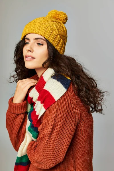 Брюнетка с вьющимися волосами в шапочке и свитере с раздетым шарфом, позирующая на сером — стоковое фото