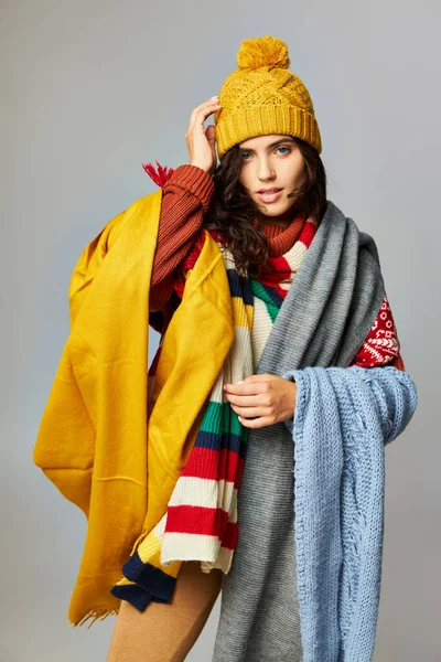 Красивая женщина с вьющимися волосами в шапочке и уютном свитере позирует с шарфами на сером фоне — стоковое фото