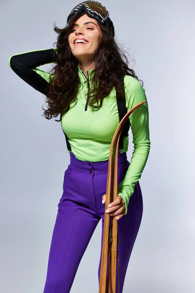 Cheerful young woman with curly hair posing in trendy active wear and holding skis on grey — Stock Photo
