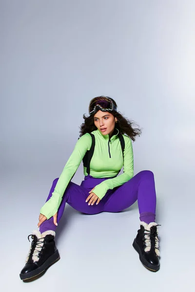 Brunette sporty woman with curly hair posing in trendy active wear while sitting on grey backdrop — Stock Photo