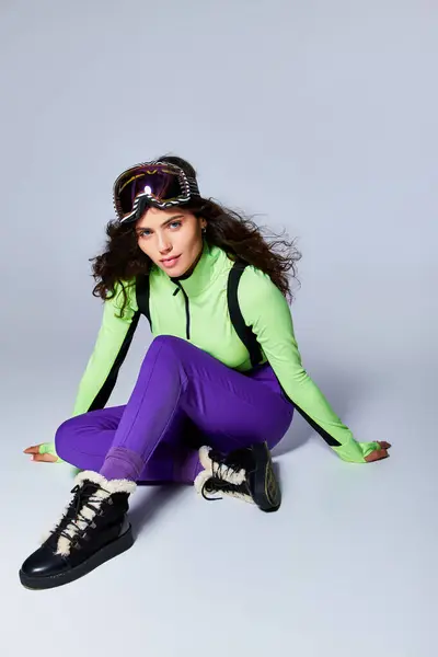 Зимний спорт, полная длина спортивной женщины с кудрявыми волосами, сидящей в активной одежде на сером фоне — стоковое фото