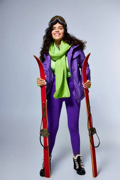 Winter sport, positive woman with curly hair posing in active wear with puffer jacket and skis — Stock Photo