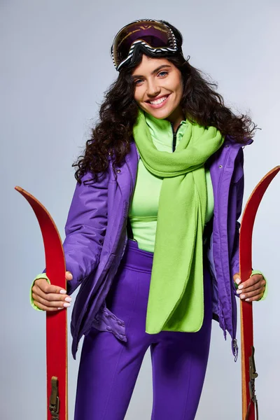 Lazer de inverno, mulher positiva com cabelo encaracolado posando em desgaste ativo com casaco de puffer e esquis — Fotografia de Stock