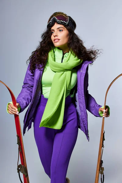 Зимний спорт, очаровательная женщина с вьющимися волосами, позирующая в активной одежде с курткой и лыжами — стоковое фото