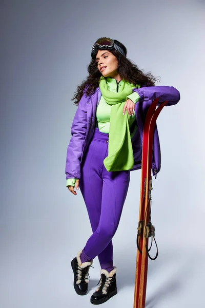 Зимняя активность, очаровательная женщина с вьющимися волосами, позирующая в активной одежде с курткой и лыжами — стоковое фото