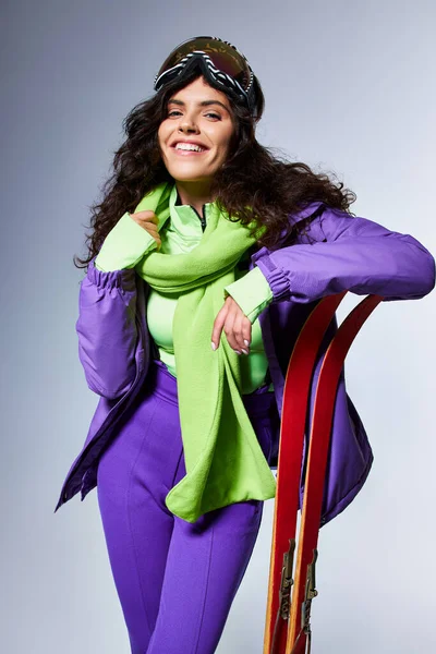 Atividade de inverno, mulher sorridente com cabelo encaracolado posando em desgaste ativo com casaco de puffer e esquis — Fotografia de Stock